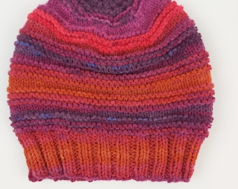 May include: A striped knitted hat in shades of purple, red, and orange. The hat has a ribbed brim and a slouchy crown.