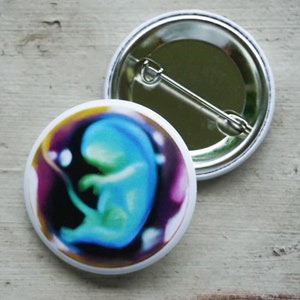 May include: Two round white buttons with a pin back. One button features a blue and green fetus illustration surrounded by purple, yellow, and black. The other button shows the pin back.