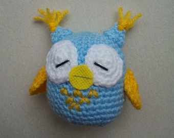 May include: A handmade crochet owl in shades of blue, white, and yellow. The owl has closed eyes, white eye circles, yellow accents on the ears and wings, and a yellow beak. The owl is a cute, handmade item.