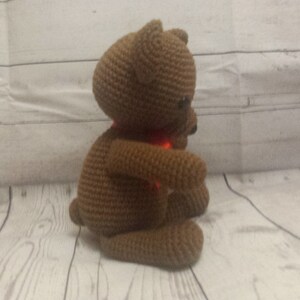 May include: A brown crocheted teddy bear with a red scarf. The bear is sitting on a white wooden surface.