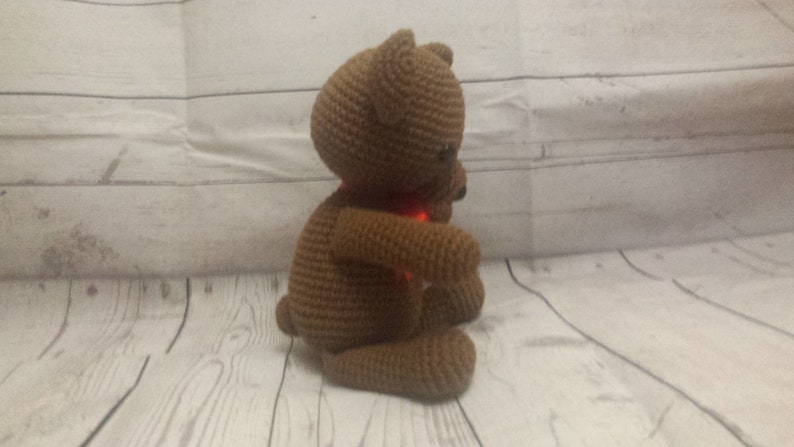 May include: A brown crocheted teddy bear with a red scarf. The bear is sitting on a white wooden surface.