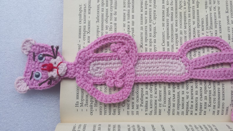 May include: A pink crocheted bookmark shaped like the Pink Panther cartoon character. The bookmark has a pink and white striped body and a pink head with black eyes and a red nose.