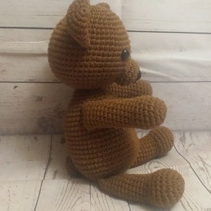 May include: A brown crocheted teddy bear sitting on a white wooden surface. The bear has black eyes and a stitched nose.