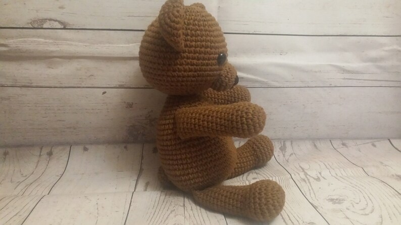 May include: A brown crocheted teddy bear sitting on a white wooden surface. The bear has black eyes and a stitched nose.