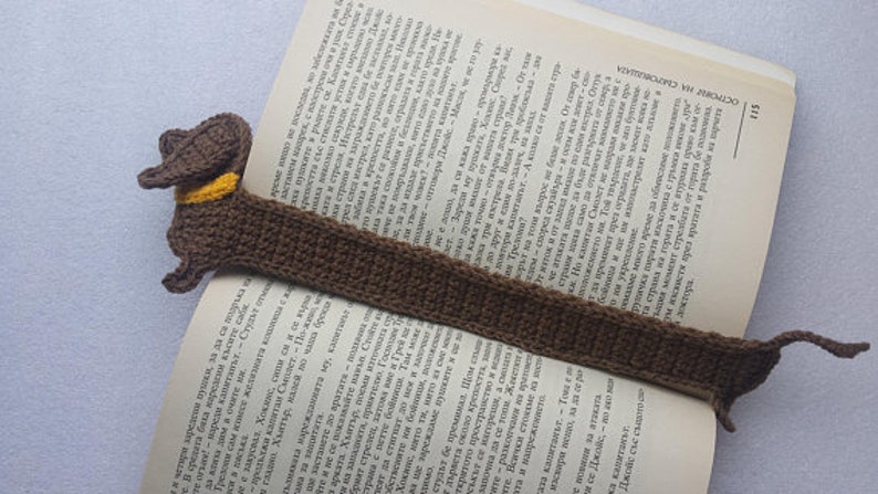 Peut inclure: Marque-page fait main en crochet en forme de chien teckel marron avec un collier jaune. Le corps du chien s'&eacute;tend le long de la page du livre ouvert, servant de marque-page. Un accessoire de lecture artisanal.