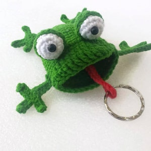 K&ouml;nnte beinhalten: Gr&uuml;ner geh&auml;kelter Frosch-Schl&uuml;sselanh&auml;nger mit gro&szlig;en wei&szlig;en Augen und einer herausgestreckten roten Zunge. Der Frosch hat einen silbernen Schl&uuml;sselring.