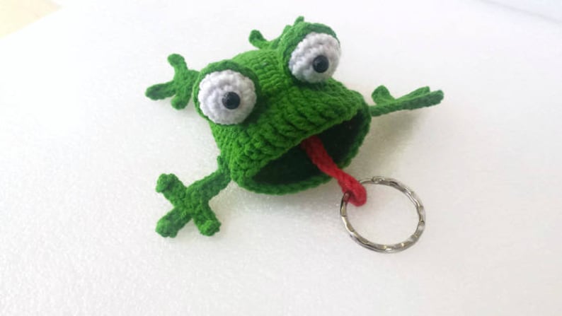 K&ouml;nnte beinhalten: Gr&uuml;ner geh&auml;kelter Frosch-Schl&uuml;sselanh&auml;nger mit gro&szlig;en wei&szlig;en Augen und einer herausgestreckten roten Zunge. Der Frosch hat einen silbernen Schl&uuml;sselring.