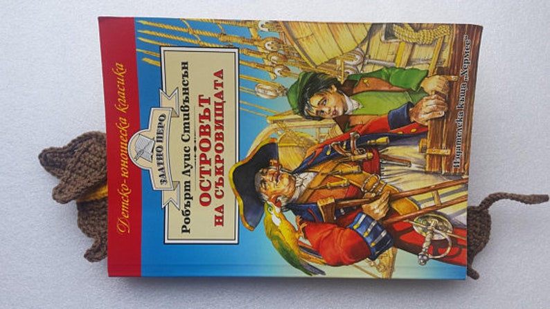 Peut inclure: Un marque-page en crochet marron en forme de souris est ins&eacute;r&eacute; dans un livre intitul&eacute; "L'&Icirc;le au tr&eacute;sor" de Robert Louis Stevenson. La couverture du livre pr&eacute;sente une illustration color&eacute;e de pirates.