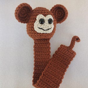 May include: A brown crocheted monkey bookmark with black eyes and a white mouth. The bookmark has a long tail.