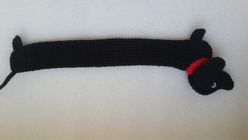 Peut inclure: Un chien teckel noir au crochet avec un collier rouge. Le chien a un long corps et de courtes pattes. La t&ecirc;te pr&eacute;sente un &oelig;il blanc et un nez noir. Fait &agrave; la main.