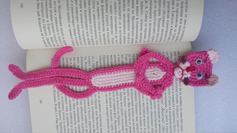 May include: A pink crocheted bookmark shaped like a cat with white accents and black eyes. The bookmark is laying on a book.