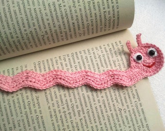 May include: A pink crocheted bookmark shaped like a worm with black eyes and a smiling mouth. The bookmark is lying on an open book.