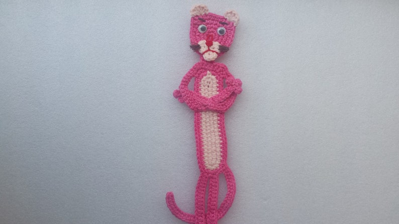 May include: Pink crocheted bookmark shaped like the Pink Panther cartoon character. The bookmark has black eyes, a black nose, and a black mustache.