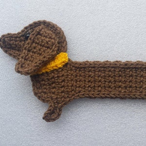 Peut inclure: Un chien teckel au crochet fait &agrave; la main, de couleur marron avec un collier jaune. Le chien est allong&eacute;, sa t&ecirc;te et son corps sont visibles. Le crochet a un aspect textur&eacute;, et le fond est gris clair.