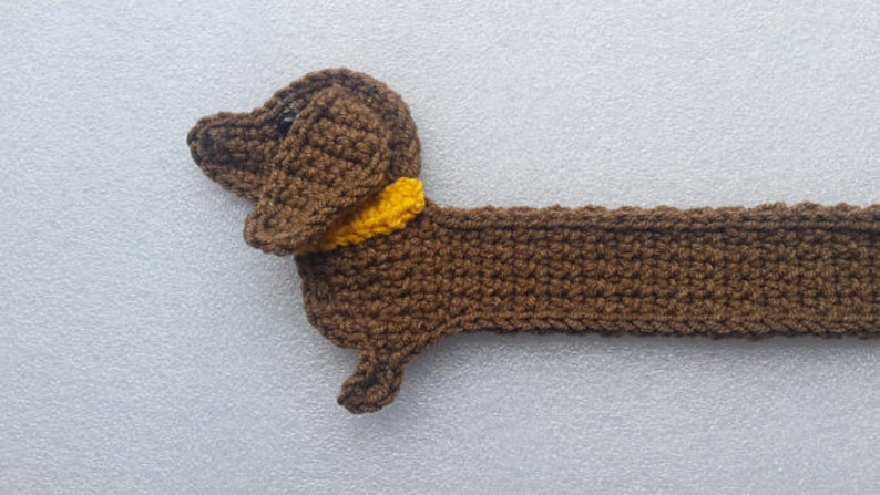 Peut inclure: Un chien teckel au crochet fait &agrave; la main, de couleur marron avec un collier jaune. Le chien est allong&eacute;, sa t&ecirc;te et son corps sont visibles. Le crochet a un aspect textur&eacute;, et le fond est gris clair.