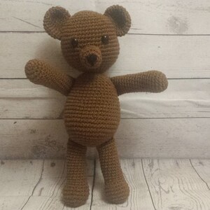 May include: A brown crocheted teddy bear with black eyes and a black nose. The bear is standing on a white wooden surface.