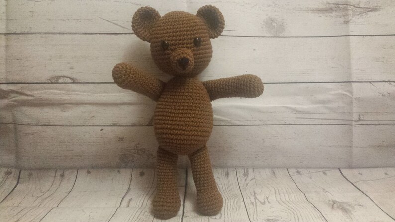 May include: A brown crocheted teddy bear with black eyes and a black nose. The bear is standing on a white wooden surface.