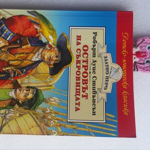 May include: A pink crocheted bookmark shaped like a cat with black eyes and a red nose. The bookmark is placed on top of a book titled "The Island of Treasure" by Robert Louis Stevenson.