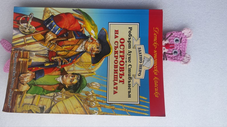 May include: A pink crocheted bookmark shaped like a cat with black eyes and a red nose. The bookmark is placed on top of a book titled "The Island of Treasure" by Robert Louis Stevenson.
