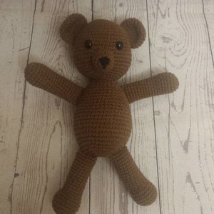 May include: A brown crocheted teddy bear with black eyes. The bear is lying on a white wooden surface.