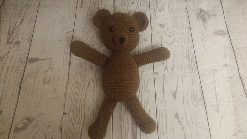 May include: A brown crocheted teddy bear with black eyes. The bear is lying on a white wooden surface.