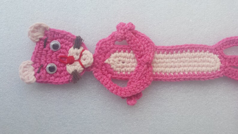 May include: Pink crocheted bookmark shaped like a cartoon panther with large eyes. The bookmark has a long tail and a pink and white body.