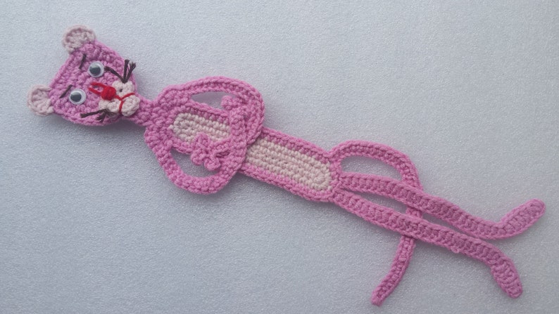 May include: Pink crocheted bookmark shaped like the Pink Panther cartoon character. The bookmark has a pink nose, black eyes, and black whiskers.