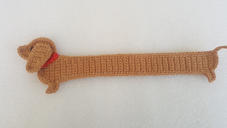 Peut inclure: Un chien teckel au crochet marron avec un collier rouge. Le chien a un long corps et de courtes pattes, et une petite queue. Le travail au crochet est d&eacute;taill&eacute;, montrant la texture du fil. Le chien mesure environ 50 cm de long.