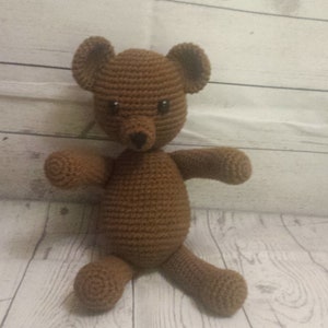 May include: A brown crocheted teddy bear with black eyes and a black nose. The bear is sitting on a light wood surface.
