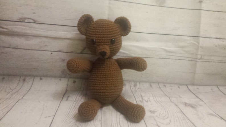 May include: A brown crocheted teddy bear with black eyes and a black nose. The bear is sitting on a light wood surface.