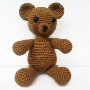 May include: A brown crocheted teddy bear with black eyes and a black nose. The bear is sitting on a white surface.