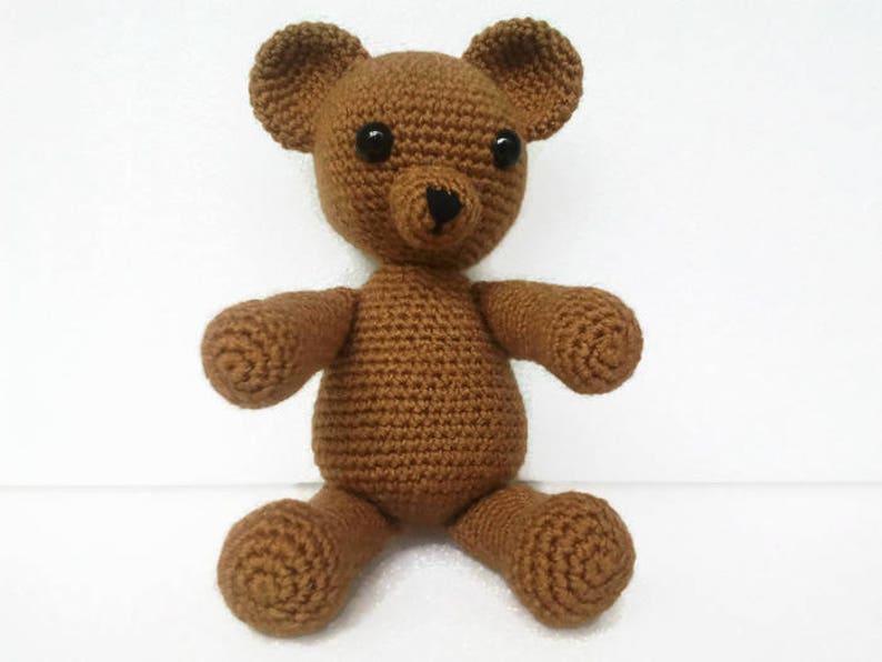 May include: A brown crocheted teddy bear with black eyes and a black nose. The bear is sitting on a white surface.