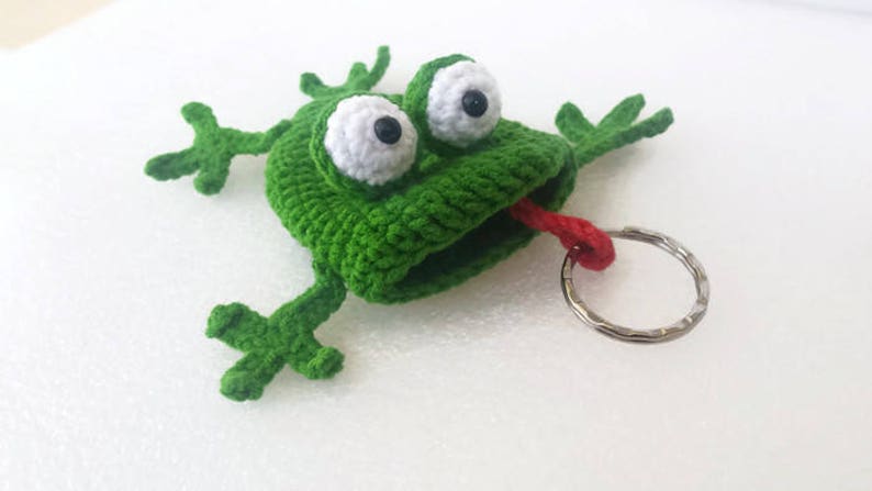 K&ouml;nnte beinhalten: Gr&uuml;ner geh&auml;kelter Frosch-Schl&uuml;sselanh&auml;nger mit gro&szlig;en wei&szlig;en Augen und einer herausgestreckten roten Zunge. Der Frosch h&auml;lt einen silbernen Schl&uuml;sselring.