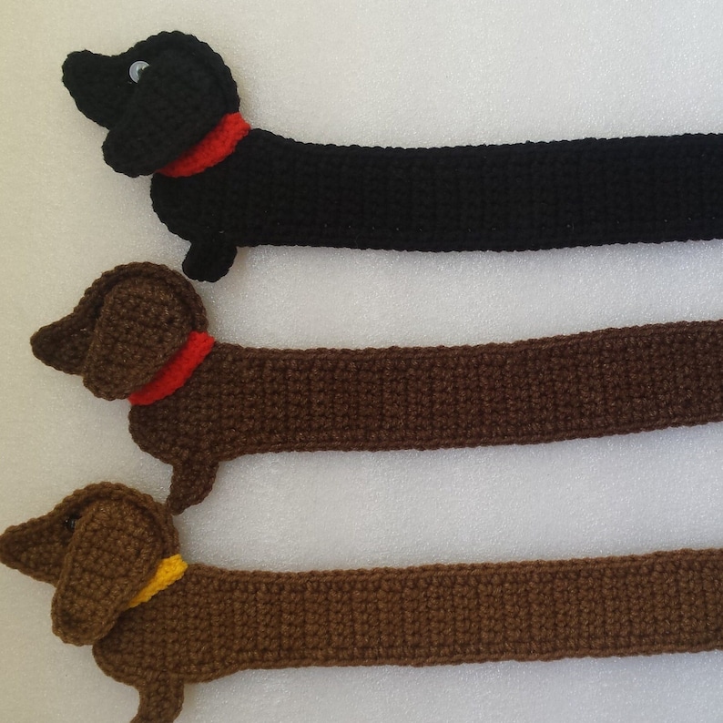 Peut inclure: Trois jouets pour chiens teckel crochet&eacute;s en noir, marron et marron clair. Chaque chien a un collier rouge ou jaune. Les chiens sont faits &agrave; la main et ont un corps long et mince et de courtes pattes.