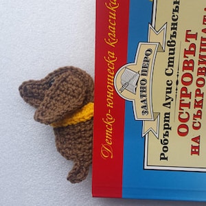 Peut inclure: Un marque-page en forme de teckel marron crochet&eacute; avec un col jaune d&eacute;passe d'un livre. Le livre a une couverture bleue avec des illustrations et du texte en langue &eacute;trang&egrave;re.