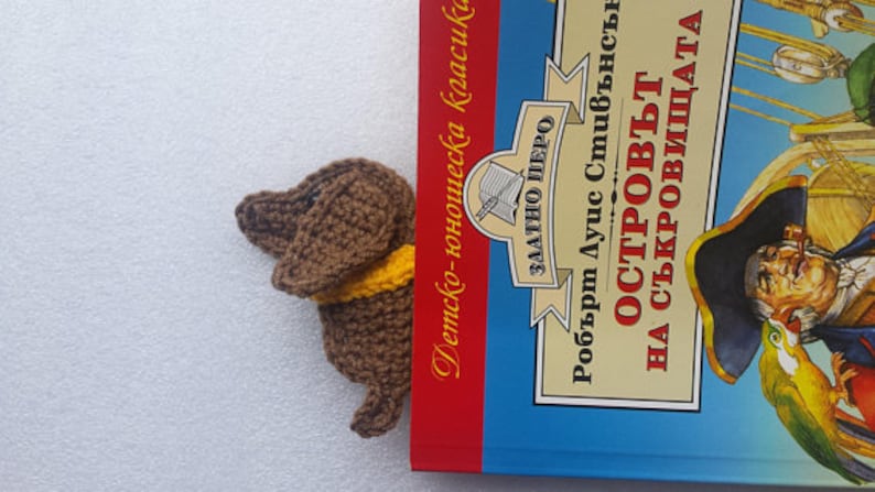 Peut inclure: Un marque-page en forme de teckel marron crochet&eacute; avec un col jaune d&eacute;passe d'un livre. Le livre a une couverture bleue avec des illustrations et du texte en langue &eacute;trang&egrave;re.