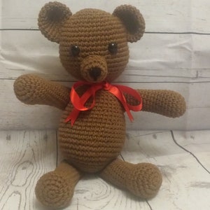 May include: A brown crocheted teddy bear with a red ribbon around its neck. The bear is sitting on a white wooden surface.