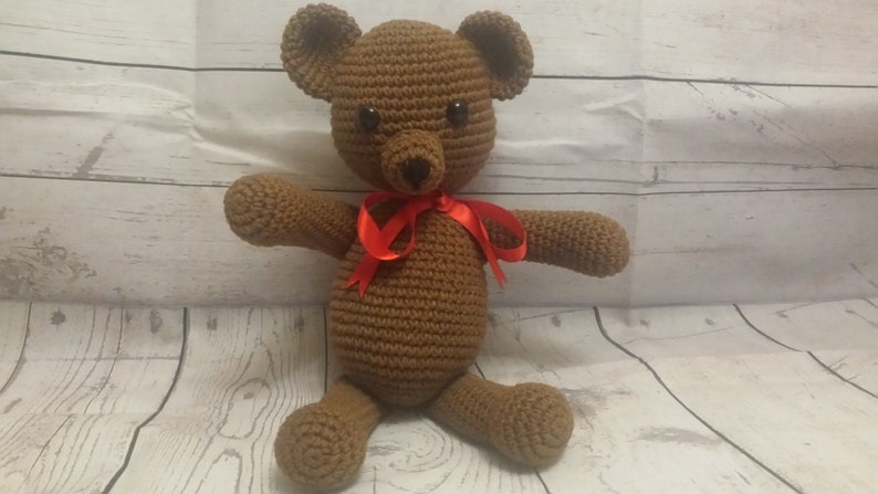 May include: A brown crocheted teddy bear with a red ribbon around its neck. The bear is sitting on a white wooden surface.