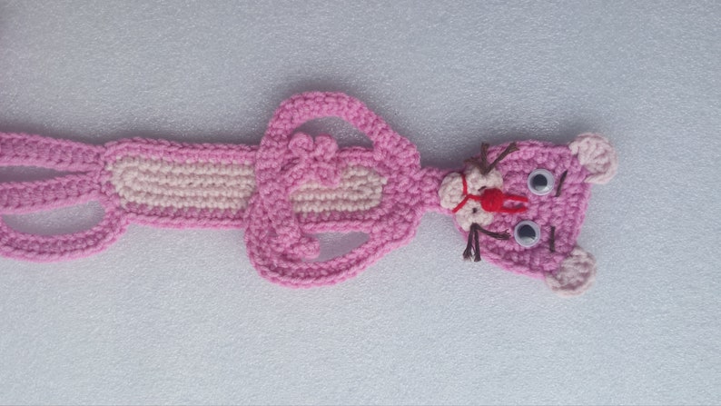 May include: Pink crocheted plush toy shaped like the Pink Panther cartoon character. The toy has a pink nose, white eyes, and brown yarn whiskers.