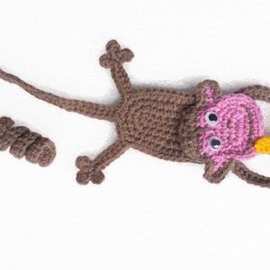 May include: A brown crocheted monkey bookmark with pink ears and a yellow banana in its hand.