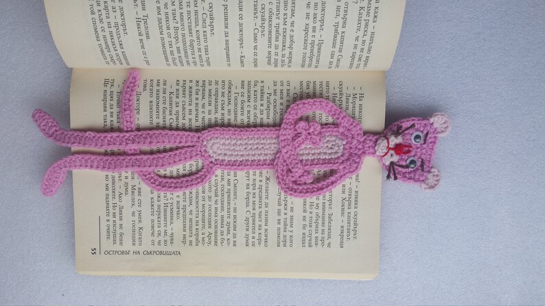 May include: A pink crocheted bookmark shaped like the Pink Panther cartoon character. The bookmark is laying on a book.