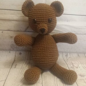 May include: A brown crocheted teddy bear with black eyes. The bear is sitting on a light wood surface.
