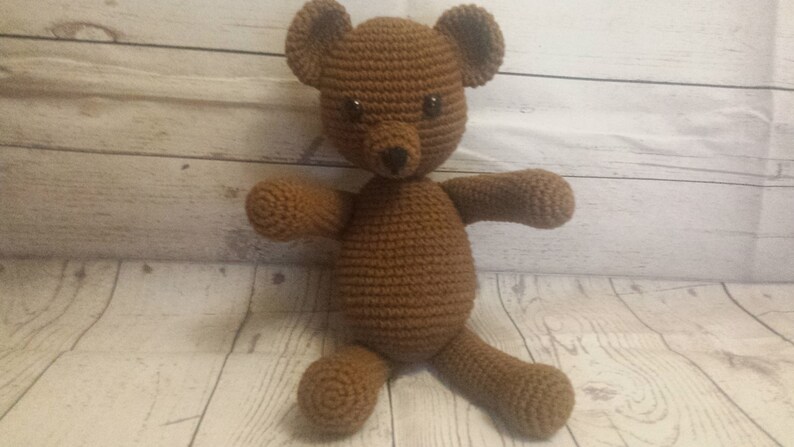 May include: A brown crocheted teddy bear with black eyes. The bear is sitting on a light wood surface.