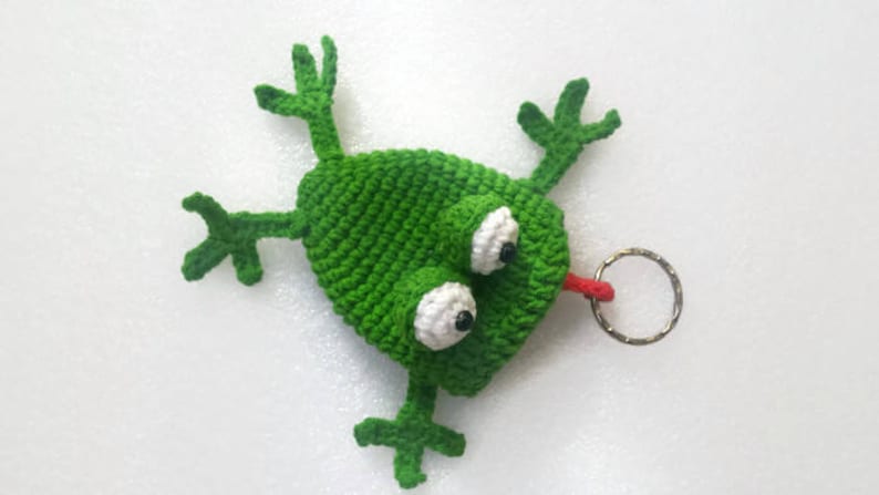 K&ouml;nnte beinhalten: Gr&uuml;ner geh&auml;kelter Frosch-Schl&uuml;sselanh&auml;nger mit gro&szlig;en wei&szlig;en Augen und einer roten Zunge. Der Frosch hat vier Beine und ist an einem silbernen Schl&uuml;sselring befestigt.
