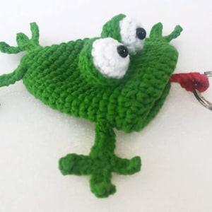 K&ouml;nnte beinhalten: Gr&uuml;ner geh&auml;kelter Frosch-Schl&uuml;sselanh&auml;nger mit gro&szlig;en wei&szlig;en Augen und einer roten Zunge. Der Frosch hat einen silbernen Schl&uuml;sselring, der an seinem Mund befestigt ist.
