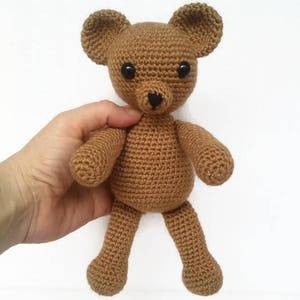 May include: A brown crocheted teddy bear with black eyes and a black nose. The bear is sitting on a white surface.