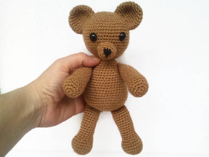 May include: A brown crocheted teddy bear with black eyes and a black nose. The bear is sitting on a white surface.