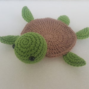 May include: A handmade crochet turtle in shades of green and brown. The turtle has a green head with black eyes, a brown shell, and green flippers. The turtle is lying on a white surface.