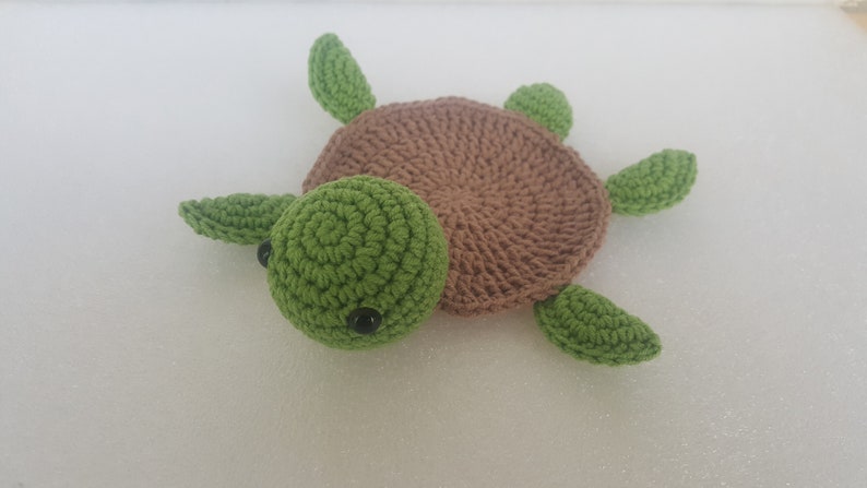 May include: A handmade crochet turtle in shades of green and brown. The turtle has a green head with black eyes, a brown shell, and green flippers. The turtle is lying on a white surface.