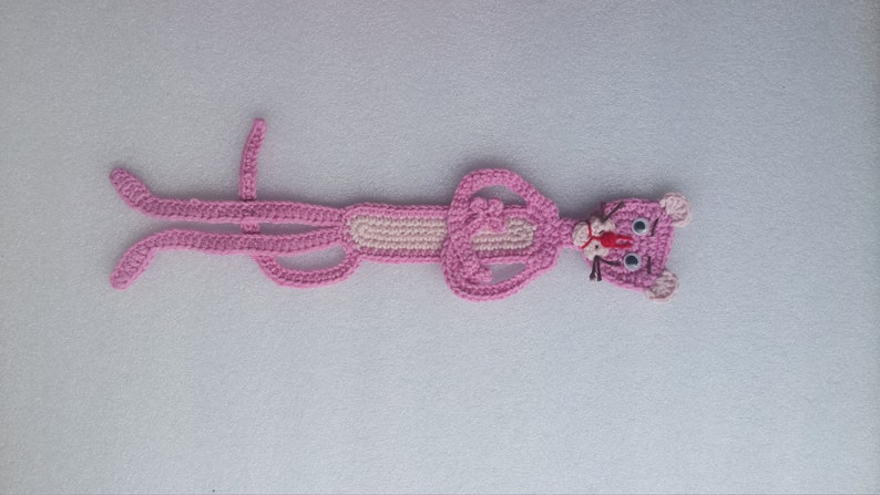 May include: Pink crocheted character of the Pink Panther lying on a white surface. The character has a pink body, white eyes, and a black nose.